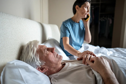 A worried caregiver talks on the phone while holding the hand of an elderly man resting in bed, suggesting a caregiving situation related to illness or medical emergency.