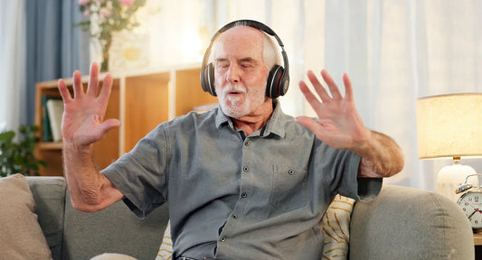 Elderly man with headphones sitting on a couch, eyes closed and hands raised, enjoying music in a cozy living room setting. The image conveys relaxation, emotion, and sensory engagement.
