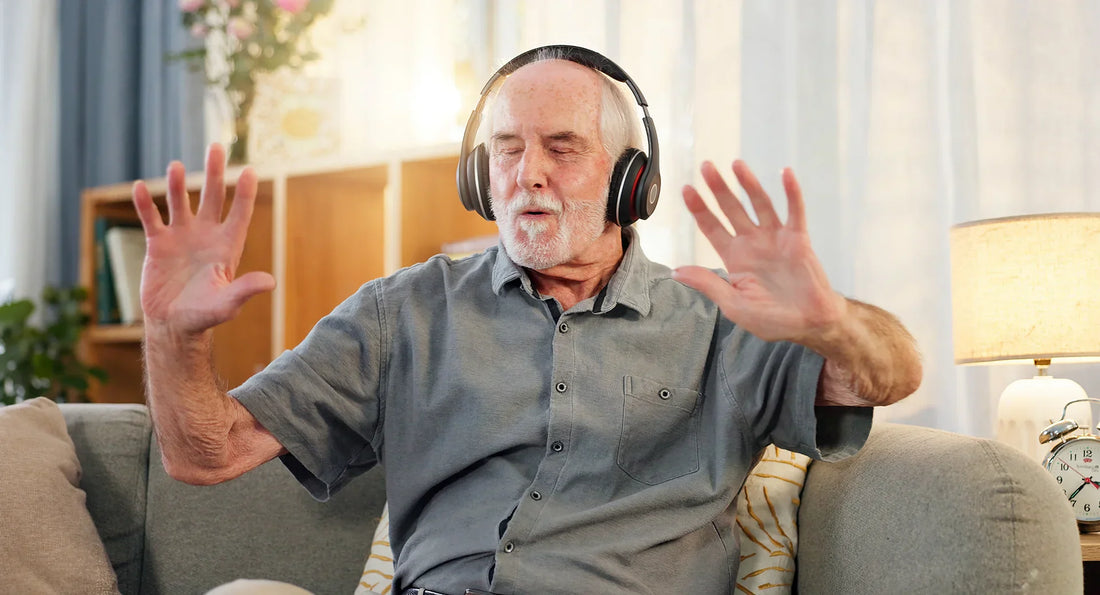 Elderly man with headphones sitting on a couch, eyes closed and hands raised, enjoying music in a cozy living room setting. The image conveys relaxation, emotion, and sensory engagement.