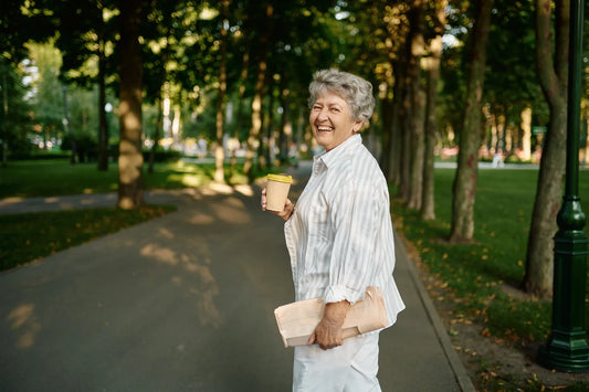 Smiling elderly woman walking through a park holding a takeaway coffee cup and a newspaper, enjoying a peaceful and sunny day that is part of her routine.