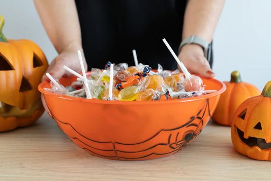 A person holding a Halloween-themed orange bowl filled with assorted lollipops and hard candies, surrounded by carved pumpkin decorations on a wooden table.